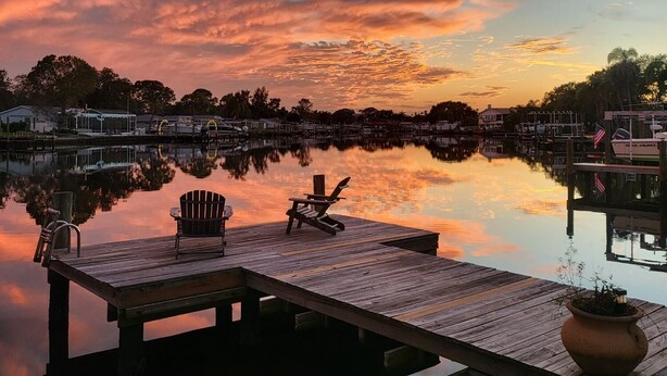 A wooden dock with two Adirondack chairs, a potted plant, and a calm body of water reflecting the sky. - Beautiful waterfront pool home 5 minutes from downtown St Pete! (St. Petersburg)