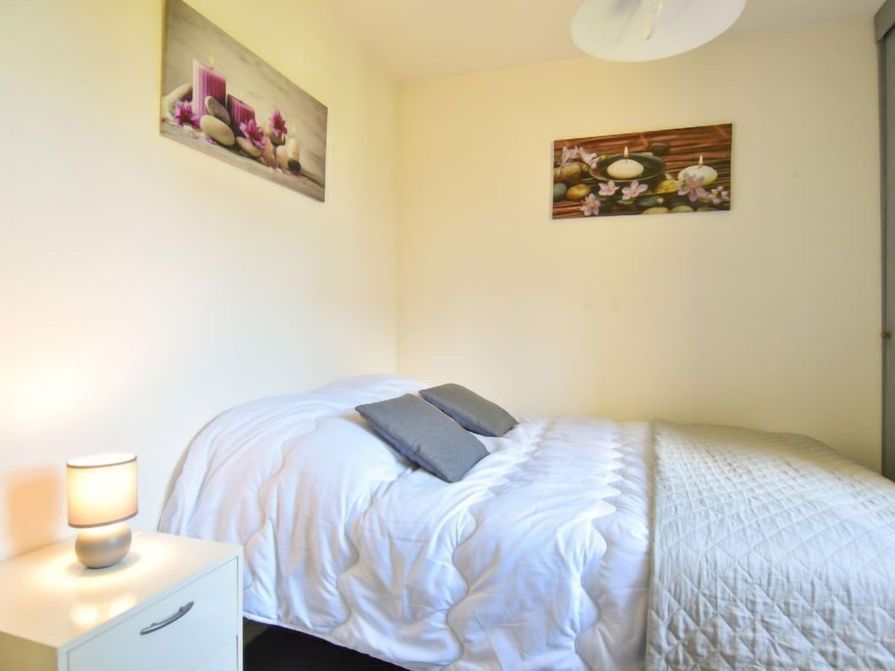 Un letto rifatto con copriletto bianco e due cuscini, una lampada da comodino e due quadri appesi raffiguranti fiori e una candela.
