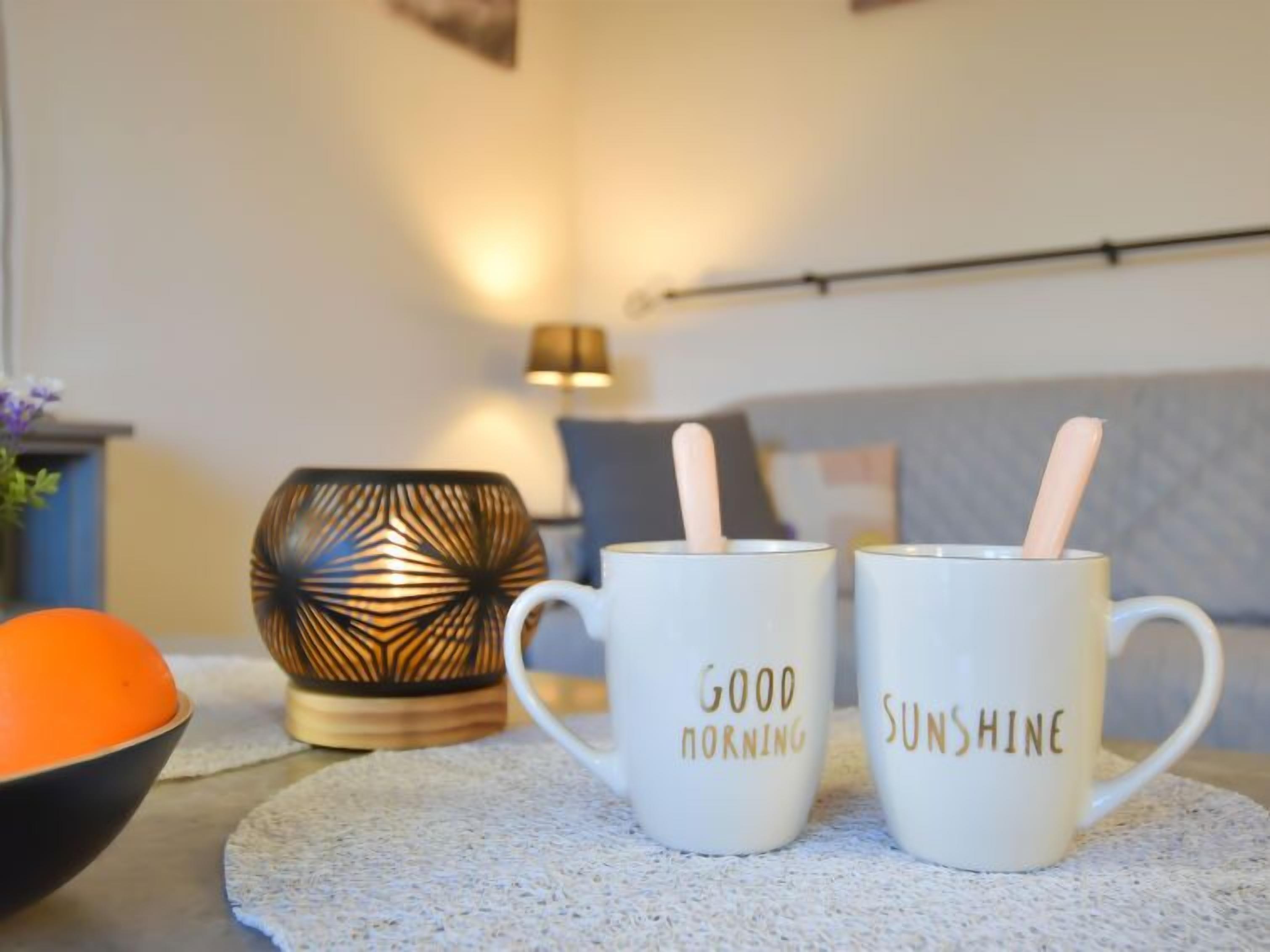 Duas canecas brancas com alças, uma com a inscrição 'GOOD MORNING' e a outra com 'SUNSHINE', sobre uma superfície texturizada.