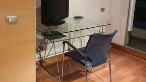 Una habitación con un escritorio de vidrio, televisión, una silla, equipo de aire acondicionado y teléfono.