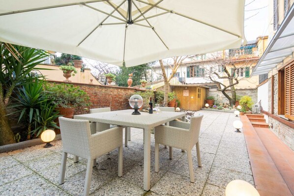Pátio externo com mesa e cadeiras, cercado por plantas em vasos e uma parede de tijolos.