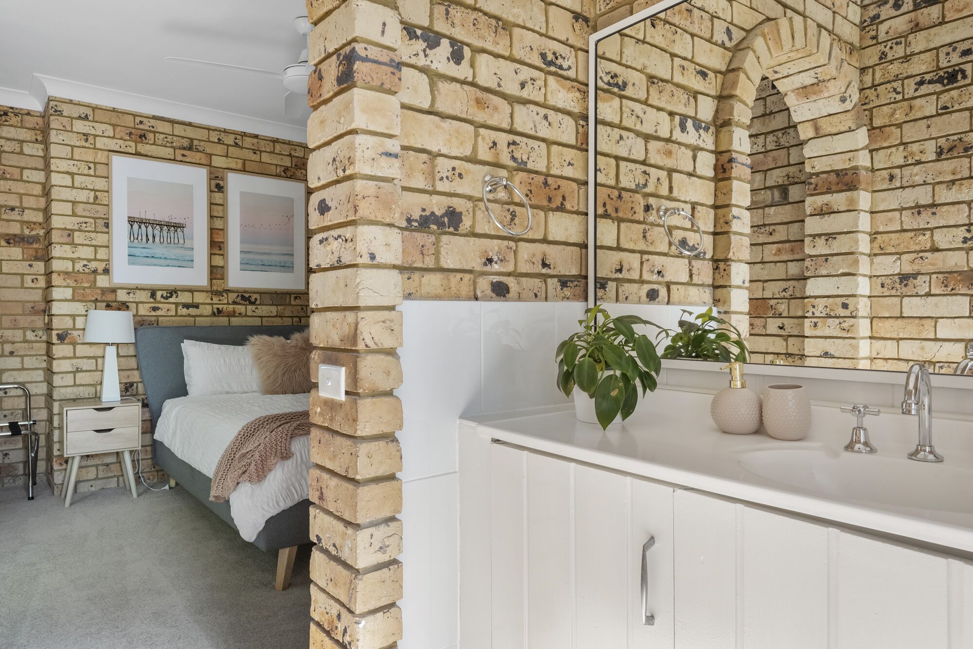A bedroom with a brick wall, a bed with a white headboard, a bedside table with a lamp, and a framed picture on the wall.