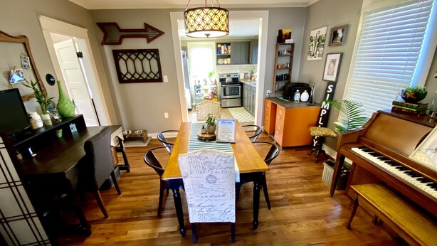 A dining area with a wooden table, chairs, a piano, and a kitchen in the background.