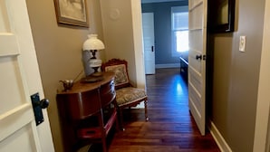 A hallway with wooden flooring, a chair, a table with a lamp, and a door leading to another room.