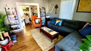 A living room with a sofa, coffee table, and bookshelves. There is a dining area in the background with a table and chairs.