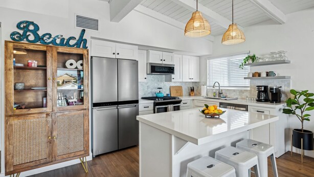 Cocina moderna con armarios blancos, refrigerador de acero inoxidable, una isla central con taburetes e iluminación colgante.