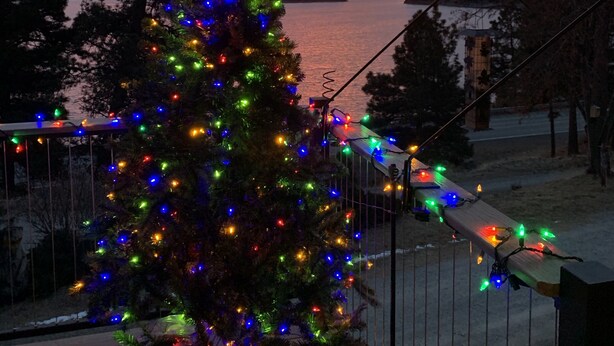 Un sapin de Noël illuminé de guirlandes colorées, placé sur une terrasse donnant sur un plan d’eau et des montagnes, au coucher du soleil.