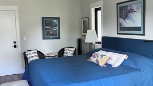 Une chambre avec un grand lit, une petite table, deux chaises, une porte blanche et deux tableaux encadrés accrochés au mur.