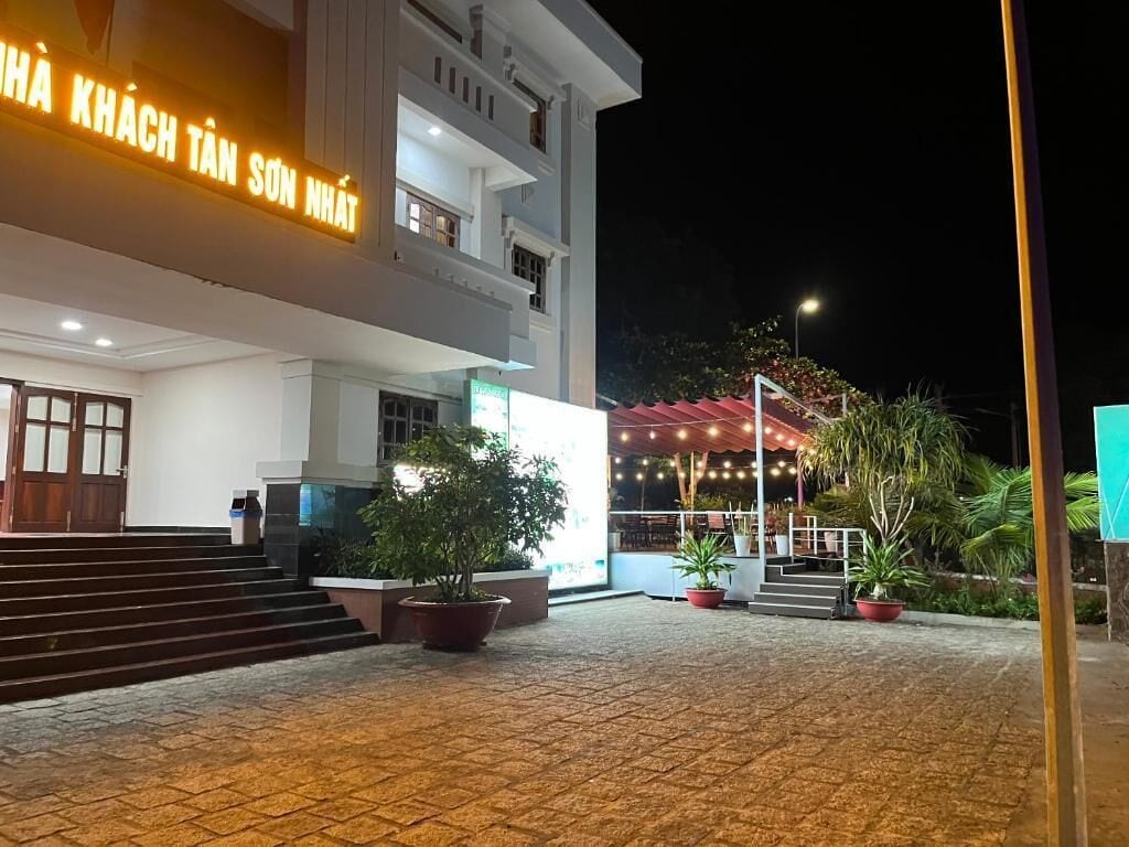 Un edificio con un letrero que indica un hotel. Hay escalones que conducen a la entrada, plantas en macetas y un espacio al aire libre cubierto con luces colgantes.
