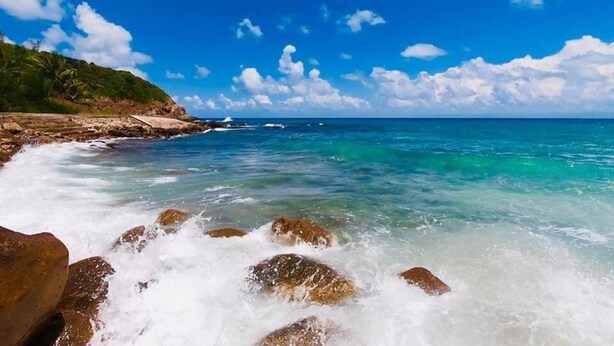 Kustlandschap met helder turquoise water, een rotsachtige kustlijn en in de verte de oceaan onder een blauwe lucht met verspreide wolken.