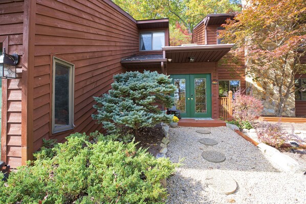 A wooden house with a green door, a window, and a small tree in the front yard.
