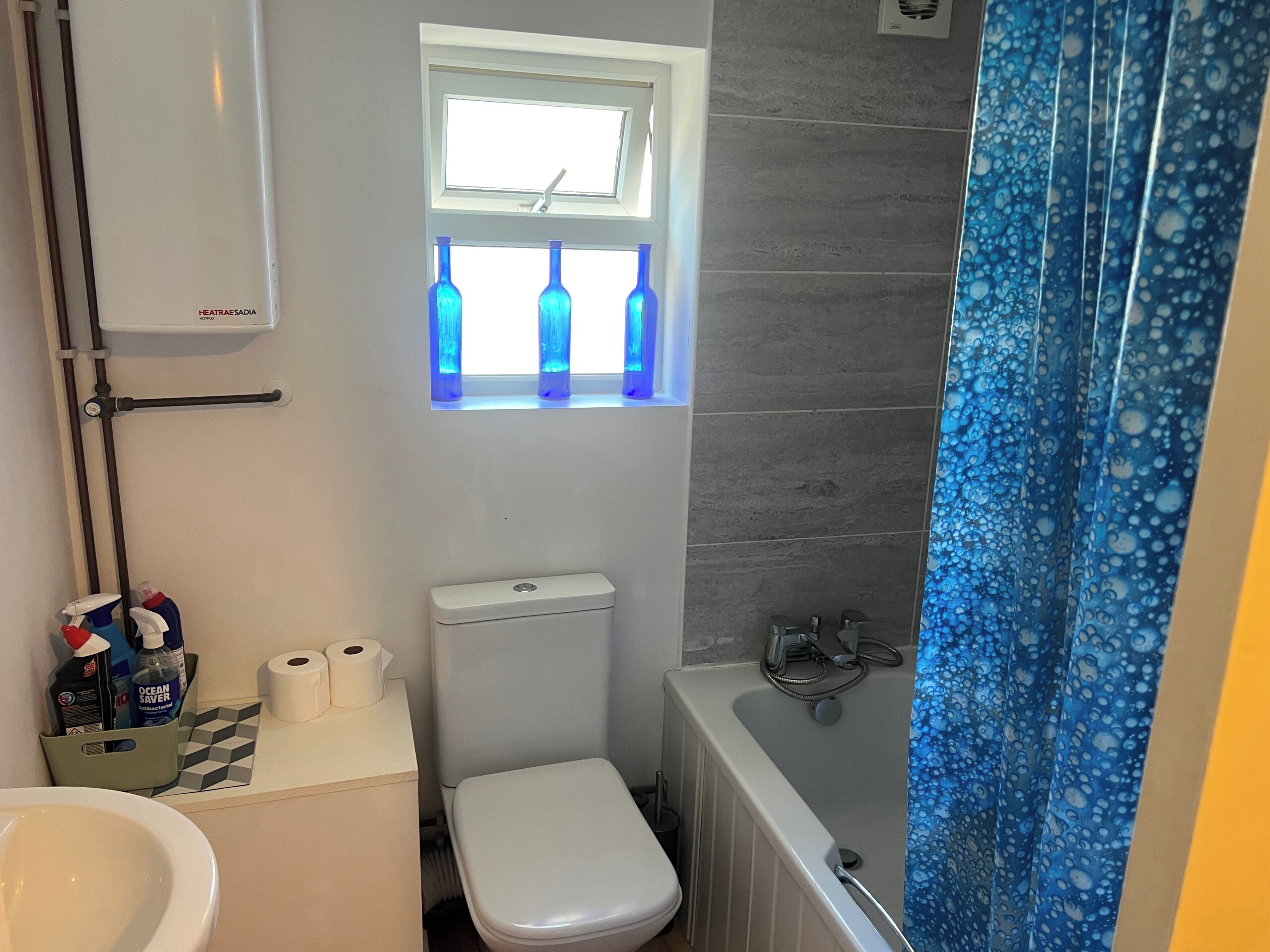 A compact bathroom with a toilet, a window with blue bottles, a radiator, and a shower curtain.