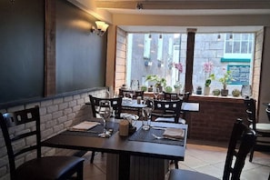 An indoor dining area with brick walls, wooden tables, and chairs. There are potted plants on a ledge and a window with a view of the street. - Charming 2 Bedroom Cornish Cottage in Mevagissey (Mevagissey, Cornwall)