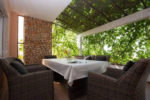 An outdoor seating area with wicker furniture and a table, covered by a pergola with a stone wall backdrop.