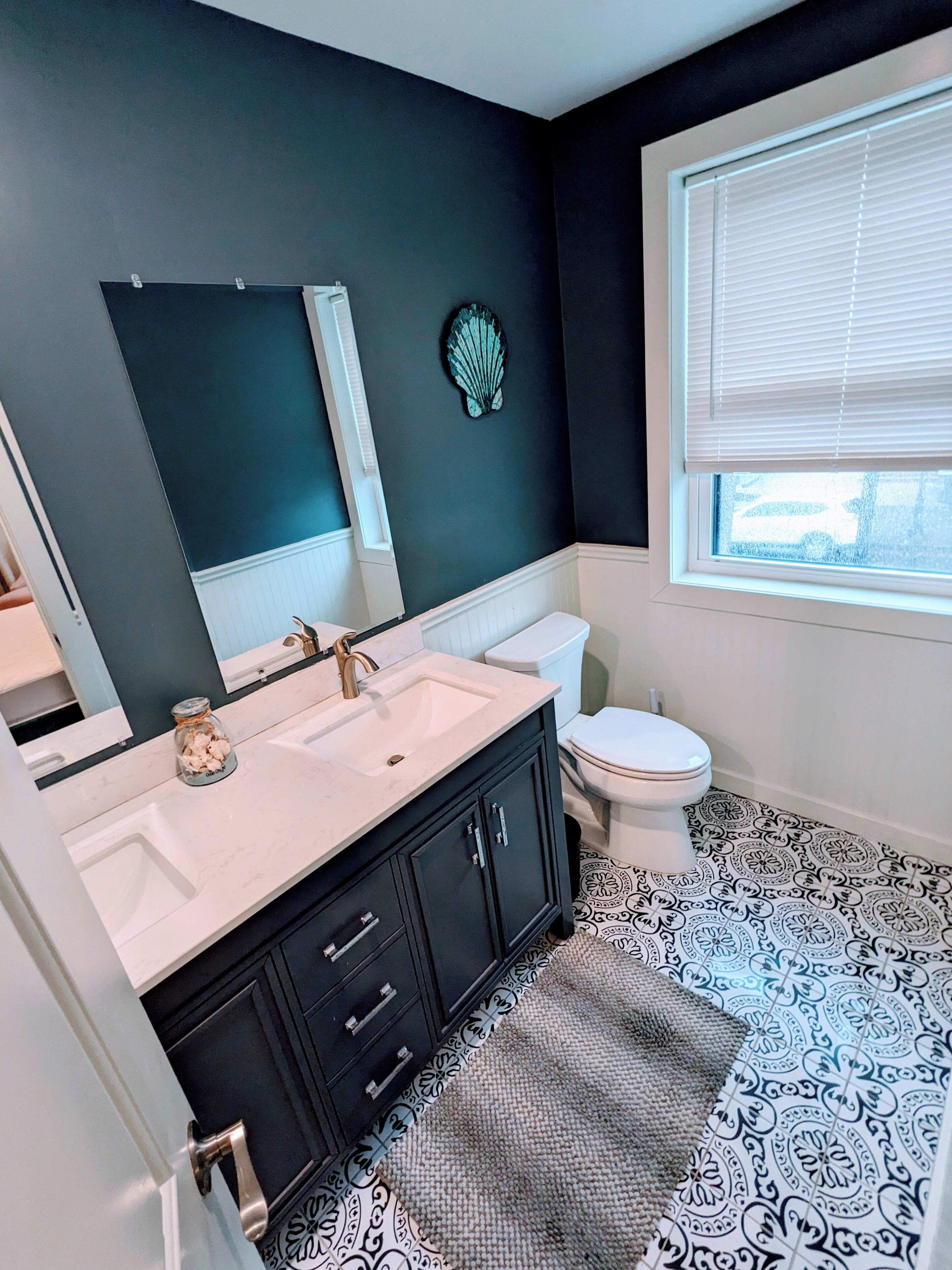 A bathroom with a black and white patterned floor, a white sink with a mirror above it, a toilet, and a window with blinds.