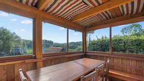 Un coin repas en bois, avec une table et des bancs, de grandes fenêtres offrant une vue sur un paysage verdoyant et des collines.