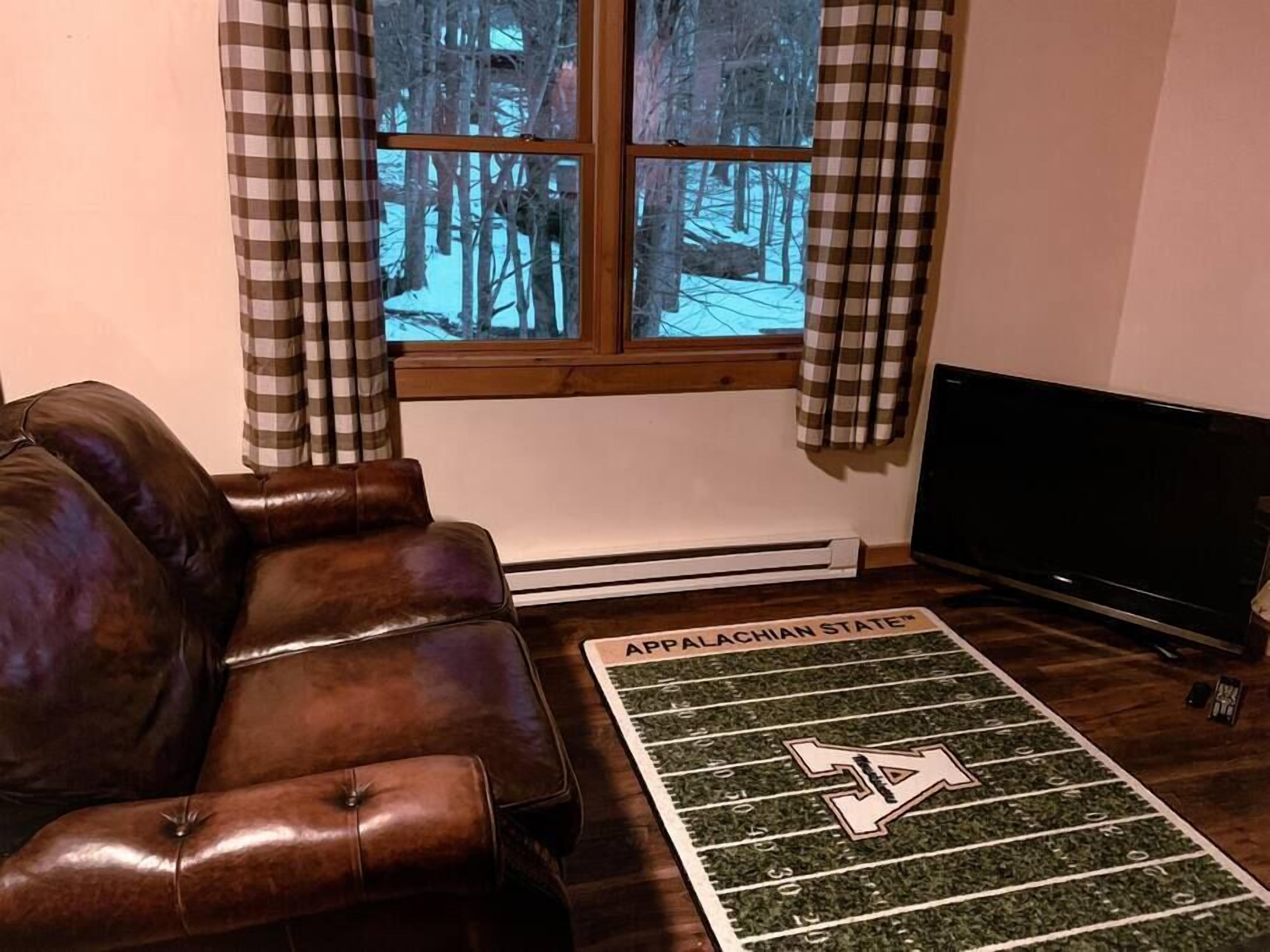 Un salon avec un canapé en cuir marron, une fenêtre ornée de rideaux à carreaux, un radiateur et un tapis représentant un terrain de football, orné du logo de l’université d’Appalachian State.