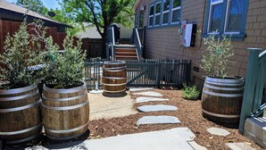 Une cour avec des tonneaux en bois utilisés comme jardinières, un chemin de pierre et un bâtiment doté d’une fenêtre et d’une porte.