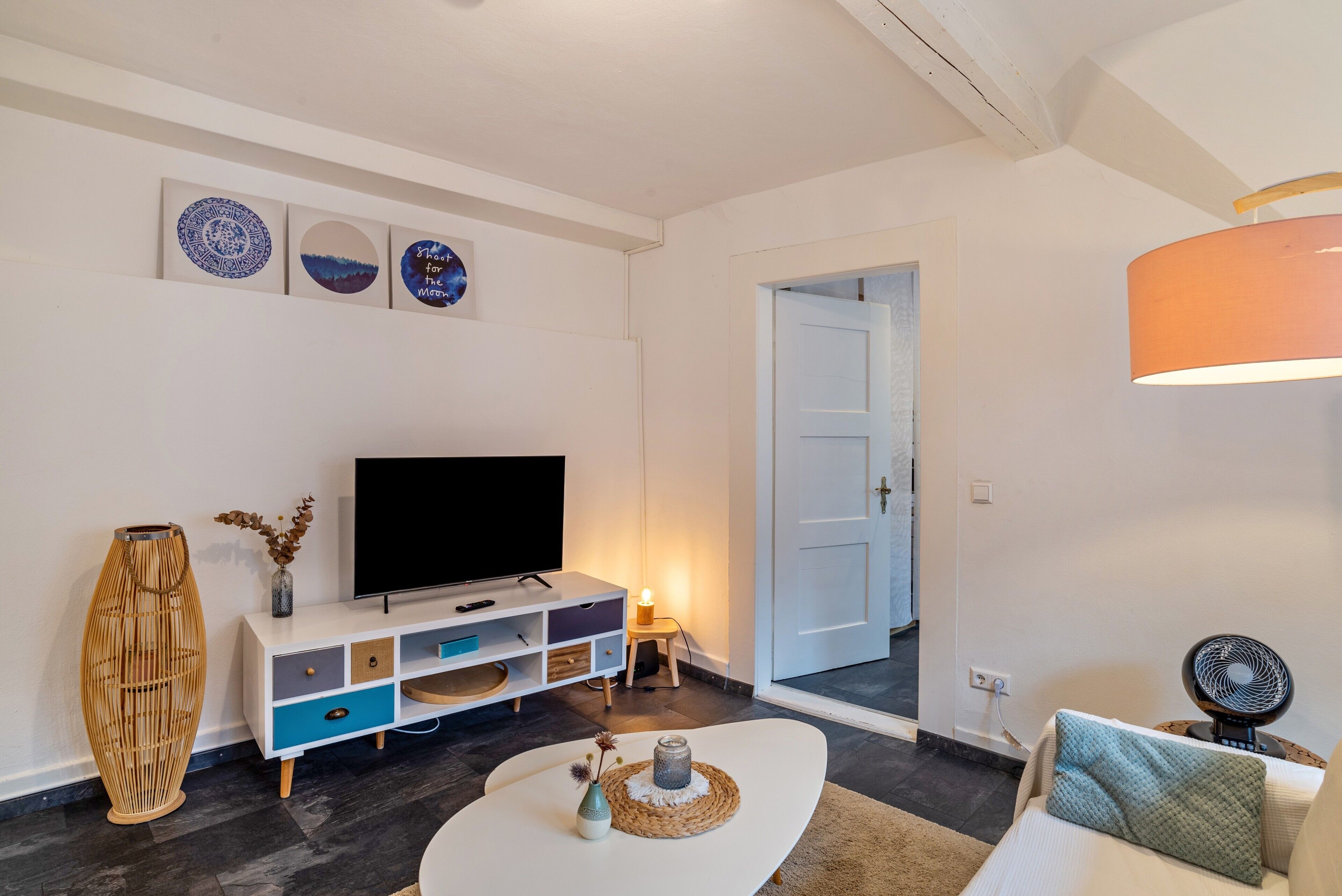 Una sala de estar moderna con un televisor de pantalla plana, una consola blanca, un florero decorativo y una mesa de centro con un posavasos tejido.
