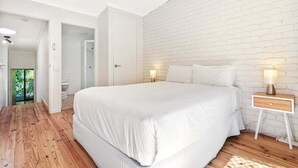 A bedroom with a large bed, white bedding, a white brick wall, and a nightstand with a lamp. - Central Noosa location in Noosa Heads (Noosa Heads)