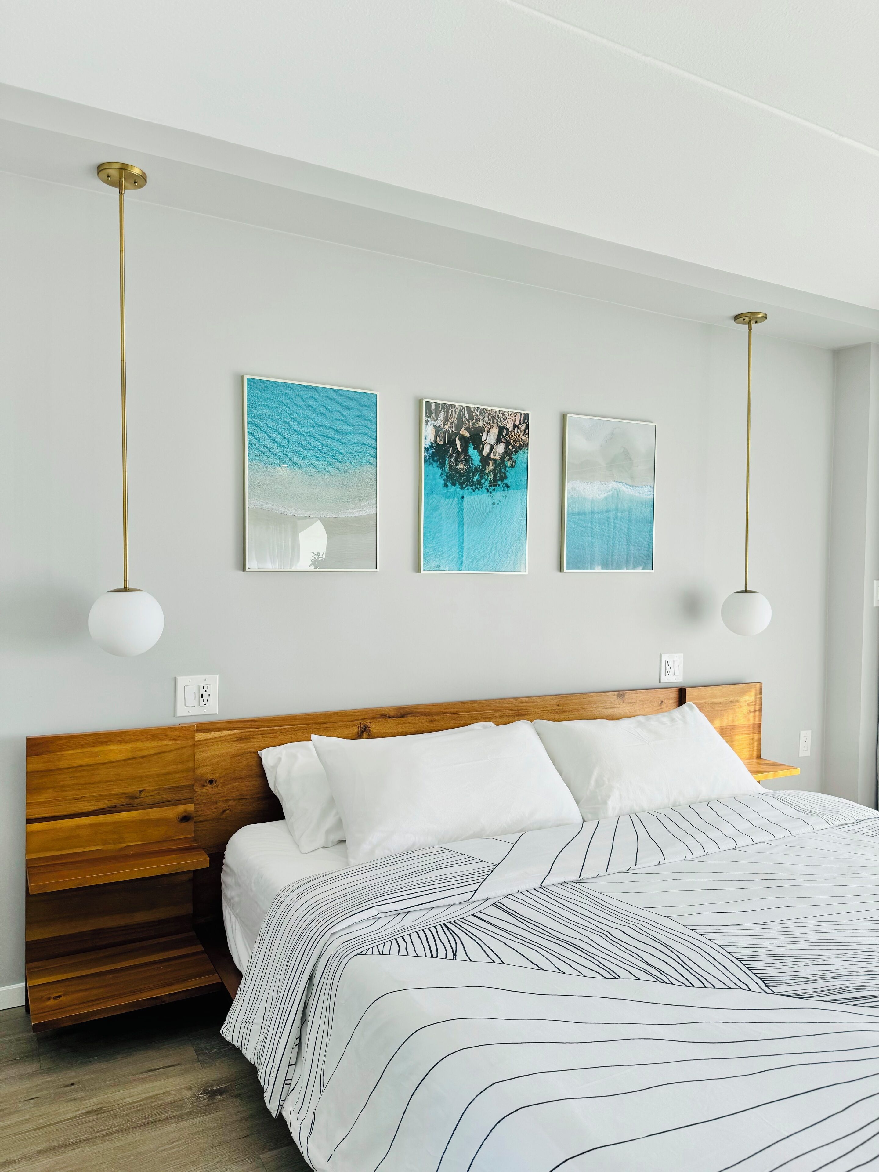 A modern bedroom with a wooden headboard, a large bed with striped bedding, and three wall-mounted artworks depicting ocean scenes.