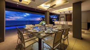 A dining area with a glass table set for six, a ceiling fan, and a view of the ocean at sunset. - The View (Koh Samui)