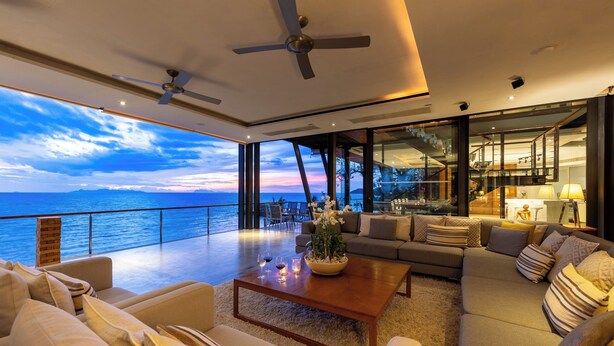 A modern living room with a large window overlooking the ocean, a ceiling fan, and a coffee table with decorative items. - The View (Koh Samui)