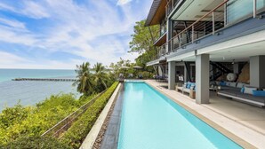 A modern poolside area with a view of the ocean, a pier, and a building with outdoor seating. - The View (Koh Samui)