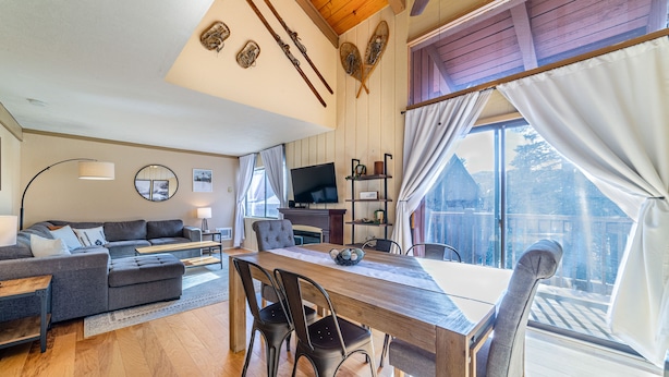 A spacious living room with a wooden dining table, grey sofa, and a TV. - ★ Summer Ready ★ Hot Tub, Summit Views, Close to Village (Mammoth Lakes)