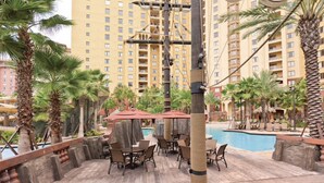 An outdoor pool area with lounge chairs and umbrellas, surrounded by palm trees and a high-rise building in the background.