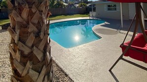 Ein Swimmingpool im Vordergrund mit einer Palme, einem roten Schaukelstuhl und einem Haus im Hintergrund.
