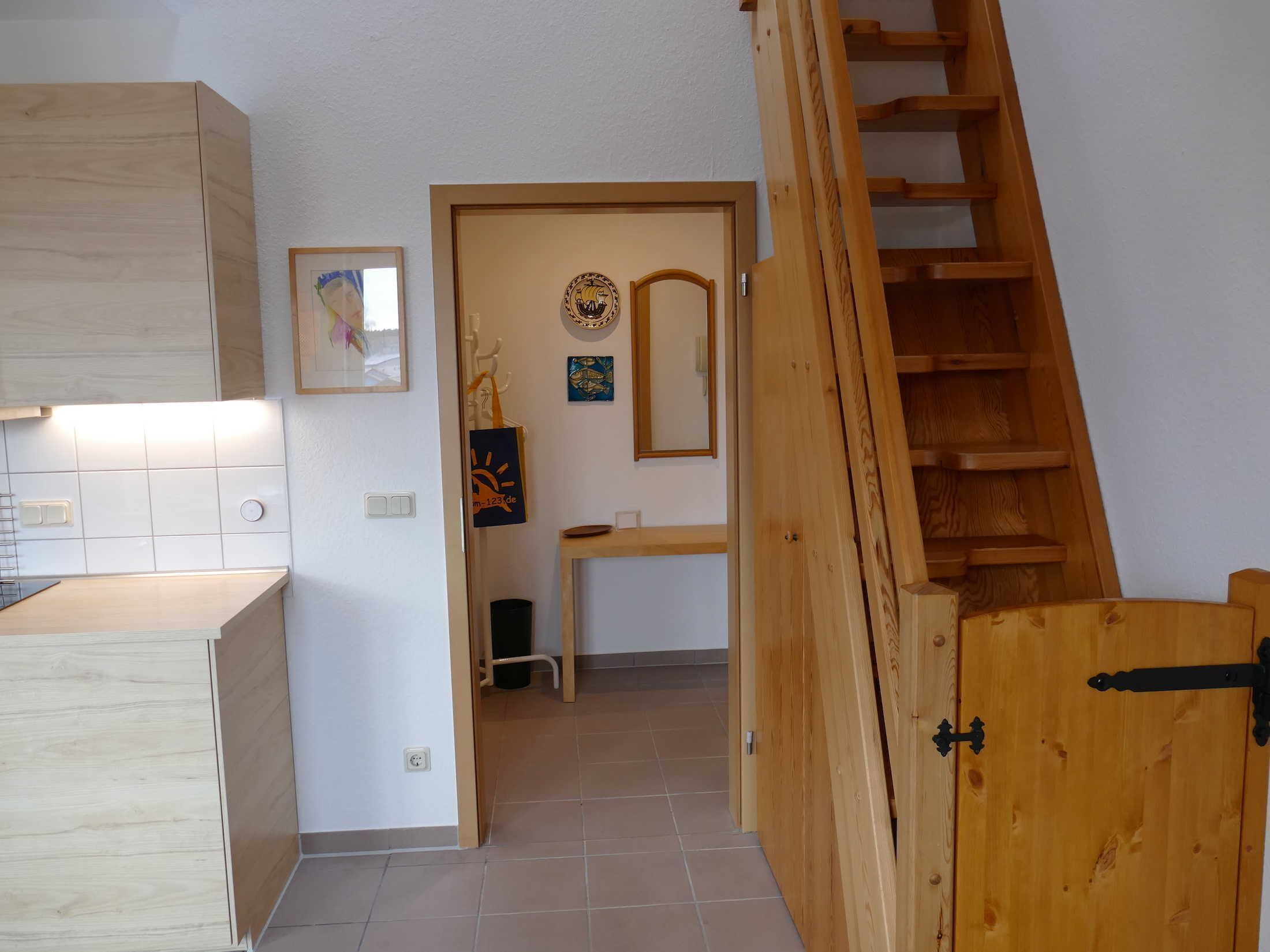 A kitchen with a wooden cabinet, a tiled backsplash, and a small table with a chair. There is a wooden staircase leading to a door with a mirror and a decorative plate on the wall.