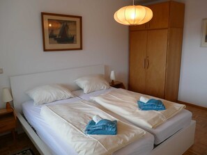 Uma cama de casal com lençóis brancos e duas toalhas, um guarda-roupa de madeira, uma mesa de cabeceira com abajur e um quadro pendurado na parede.