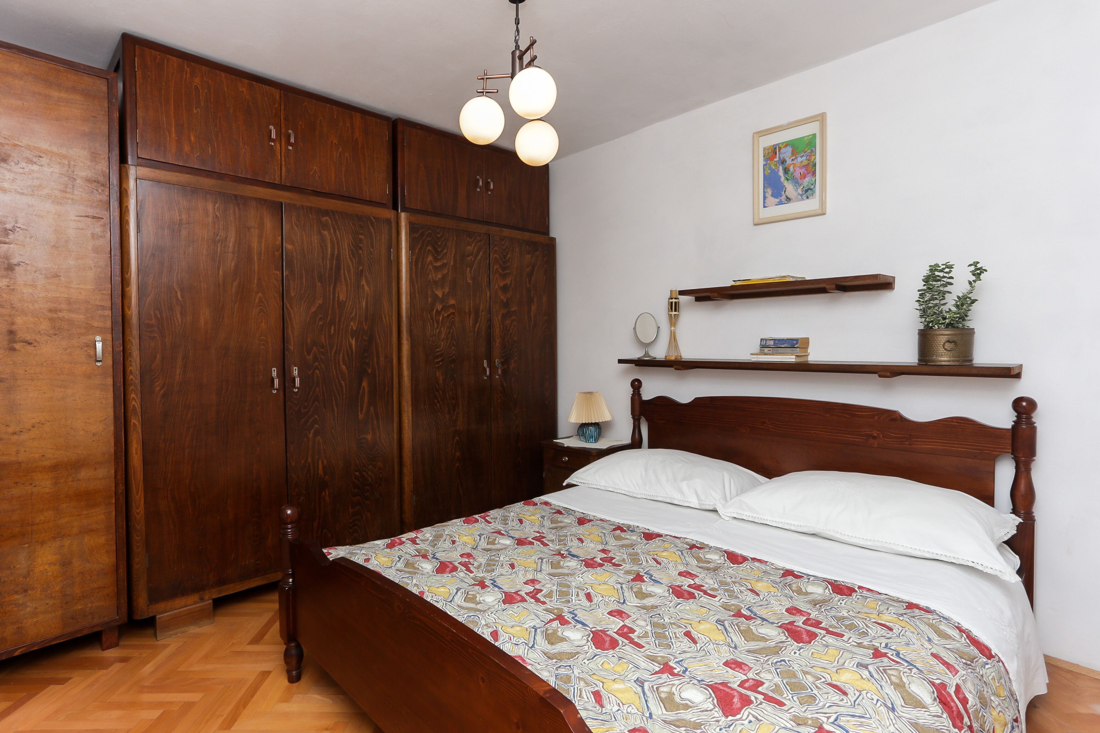 A bedroom with a wooden bed, a wardrobe, a bedside table with a lamp, and a shelf with a plant.