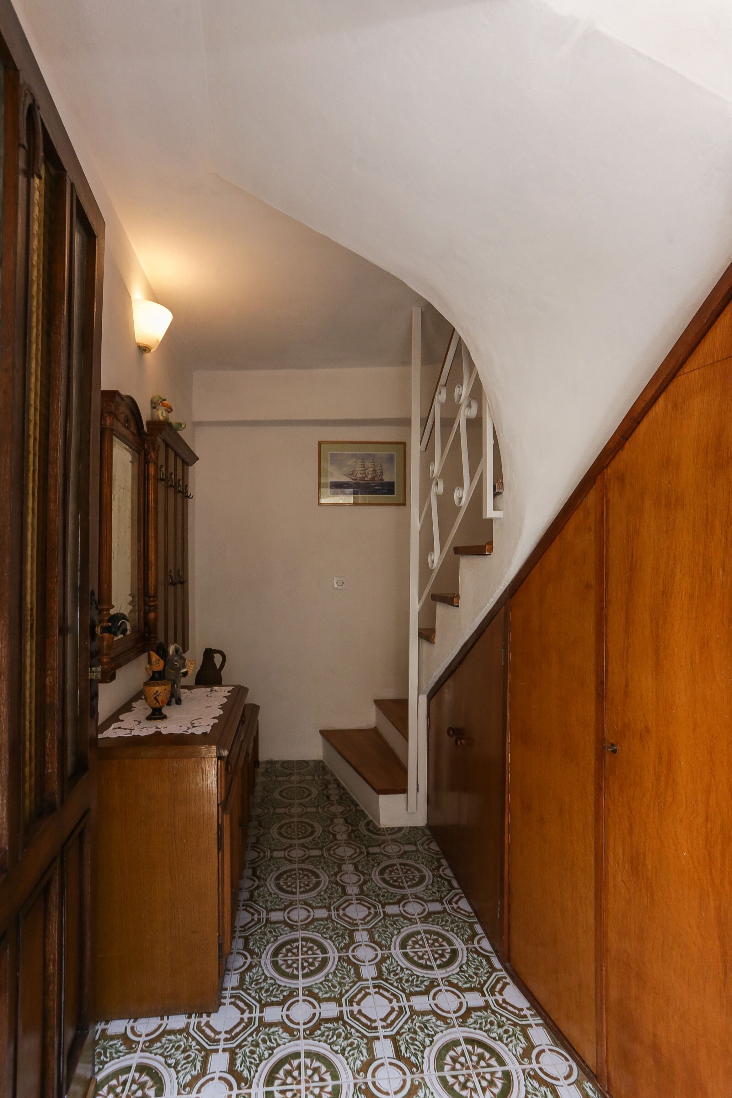 A narrow hallway with patterned floor tiles, wooden furniture, and a staircase with white railings.