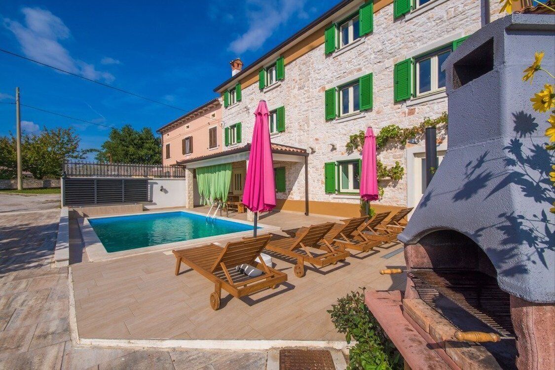 A stone building with a swimming pool, wooden lounge chairs, and a barbecue grill.