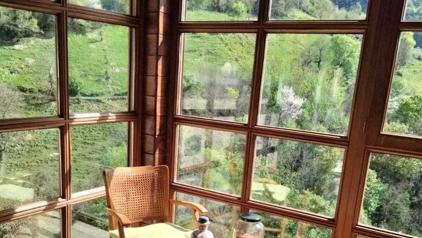 Une pièce dotée de grandes fenêtres donnant sur un paysage verdoyant. On y trouve une chaise en bois et une petite table sur laquelle sont posés une bouteille et un bocal.