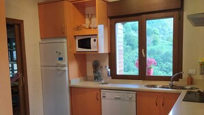 Une cuisine compacte avec des meubles en bois, un réfrigérateur blanc, un four à micro-ondes et un lave-vaisselle. Une fenêtre offre une vue sur la verdure environnante.