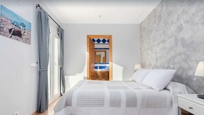 A bedroom with a bed, a nightstand, a lamp, a window with curtains, a door with a mirror, and a wall with a textured pattern. - Beach house for 6 persons, with pool and garden, in Playas de Muro, Mallorca (Muro)