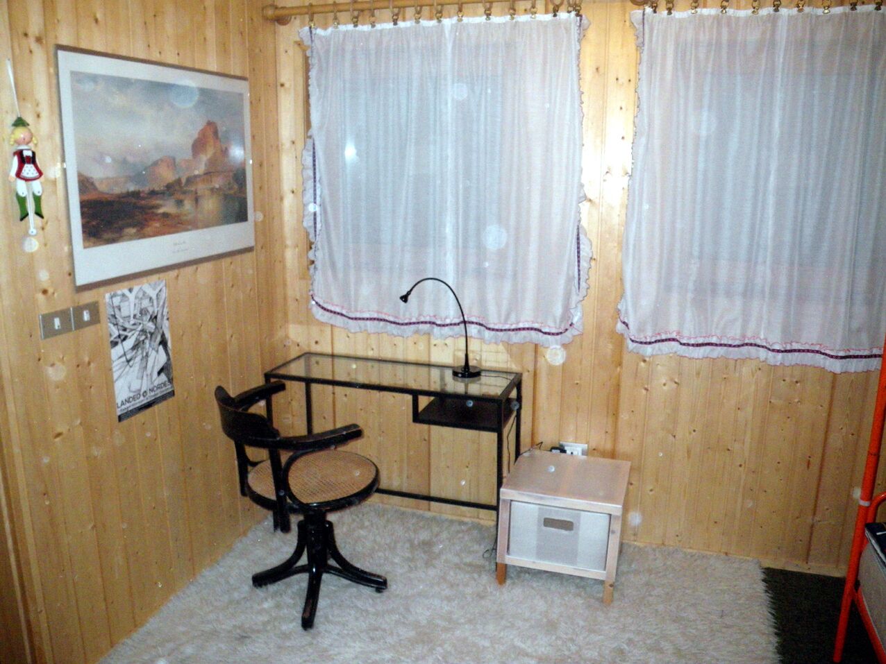 Une petite pièce aux murs en bois, un bureau avec une chaise, deux fenêtres avec des rideaux, un tableau encadré au mur et un petit meuble.