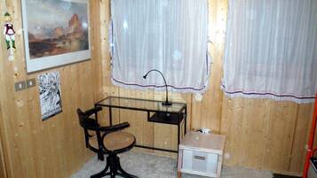 Une petite pièce aux murs en bois, un bureau avec une chaise, deux fenêtres avec des rideaux, un tableau encadré au mur et un petit meuble.