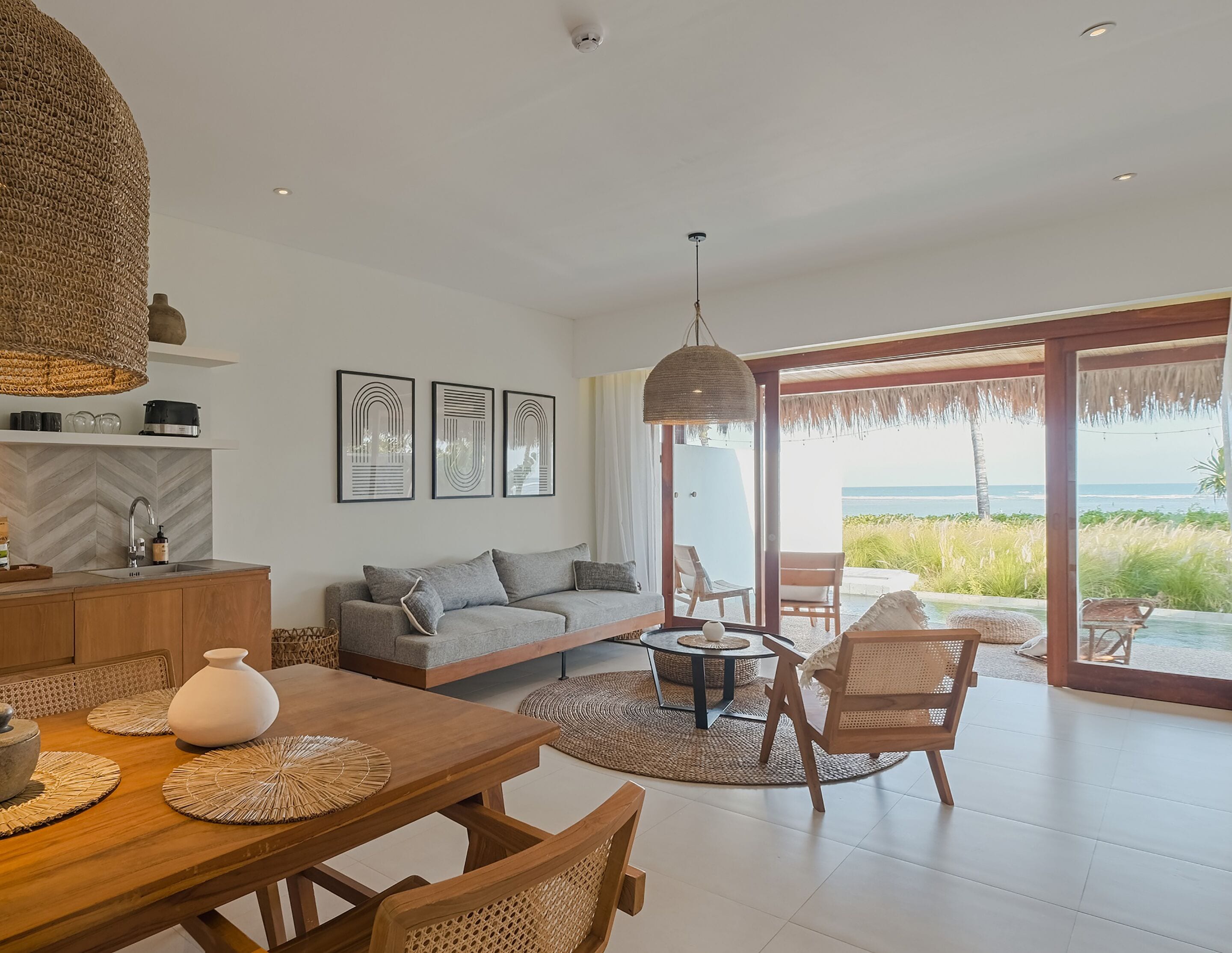 A modern living room with a sofa, dining table, and chairs, a kitchen area, and a large glass door leading to a balcony with a view of the ocean.