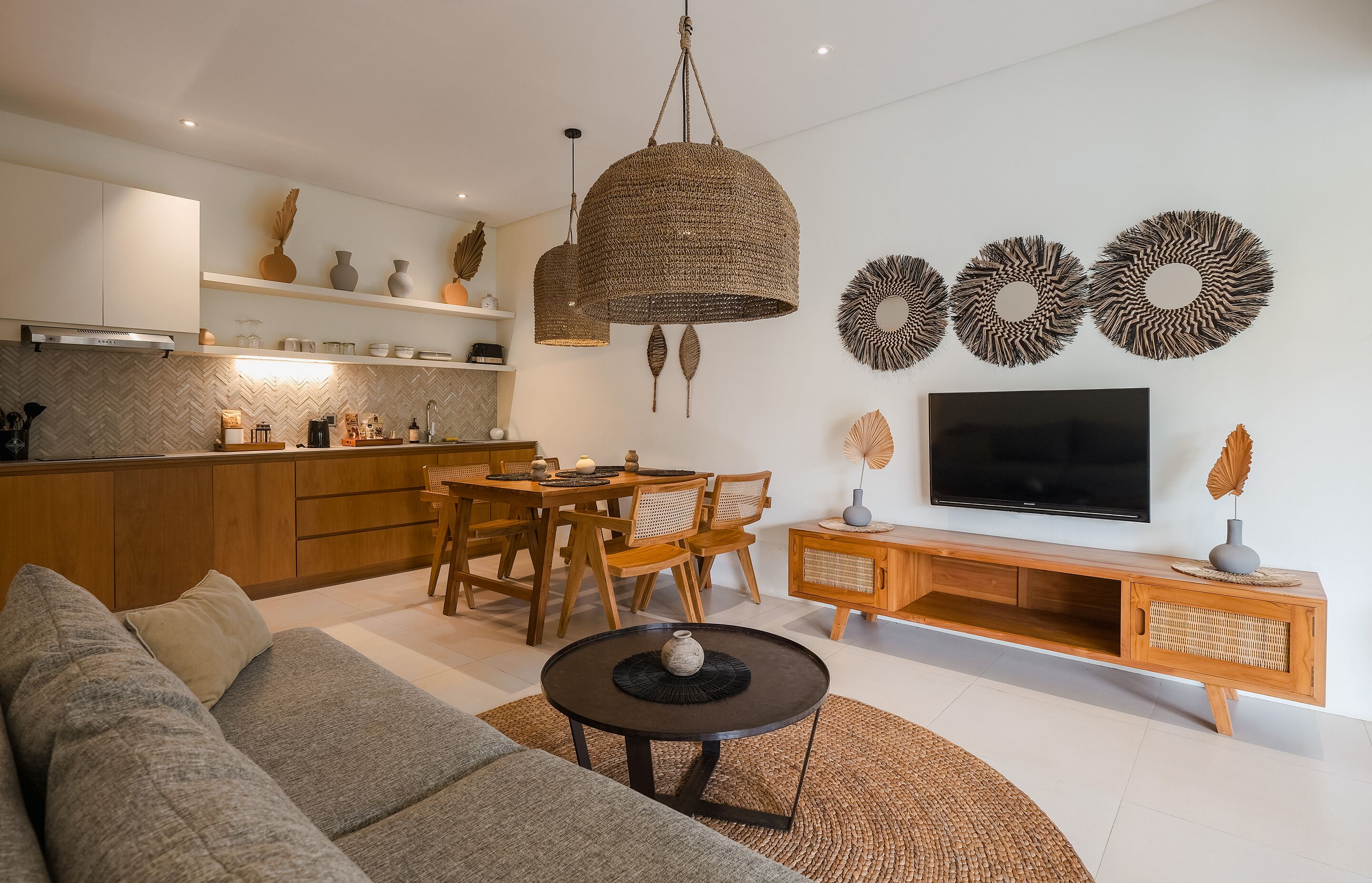 A modern living room with a sofa, coffee table, and TV stand. There is a dining area with a table and chairs. The kitchen is visible with cabinets and a stove.