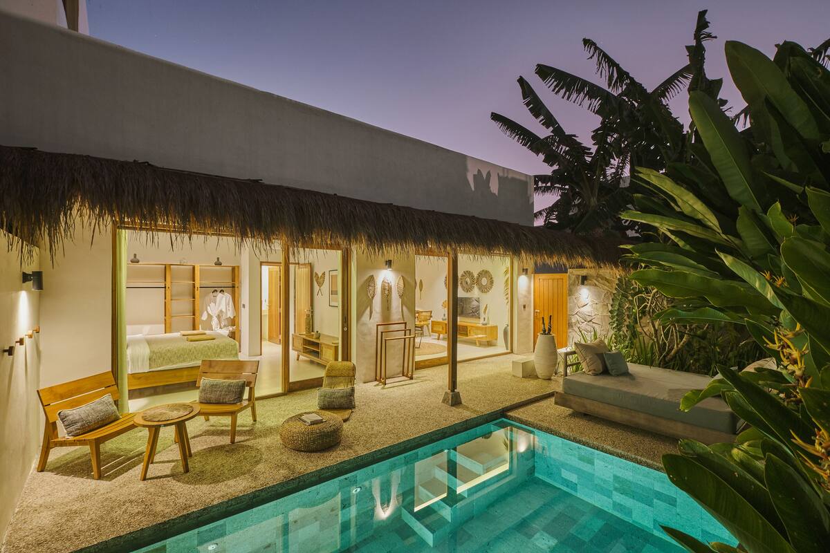 A pool area with a thatched roof, wooden furniture, and a stone wall.
