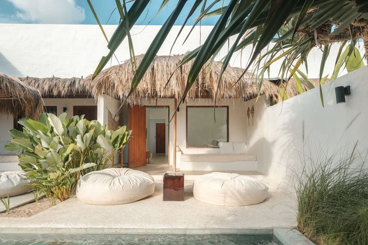 A pool area with a thatched roof, white walls, and a seating area with cushions.