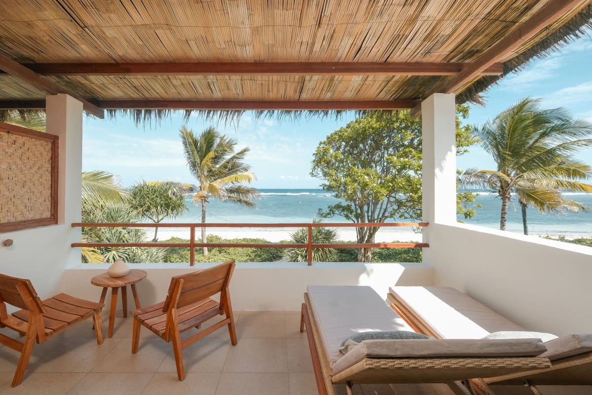 A balcony with a wooden ceiling, two wooden chairs, a small round table, and a bed with a white mattress.