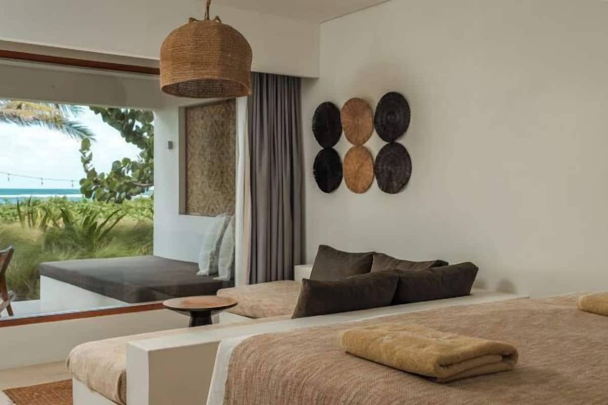 A modern bedroom with a large bed, a small seating area, a woven light fixture, and a view of greenery outside.