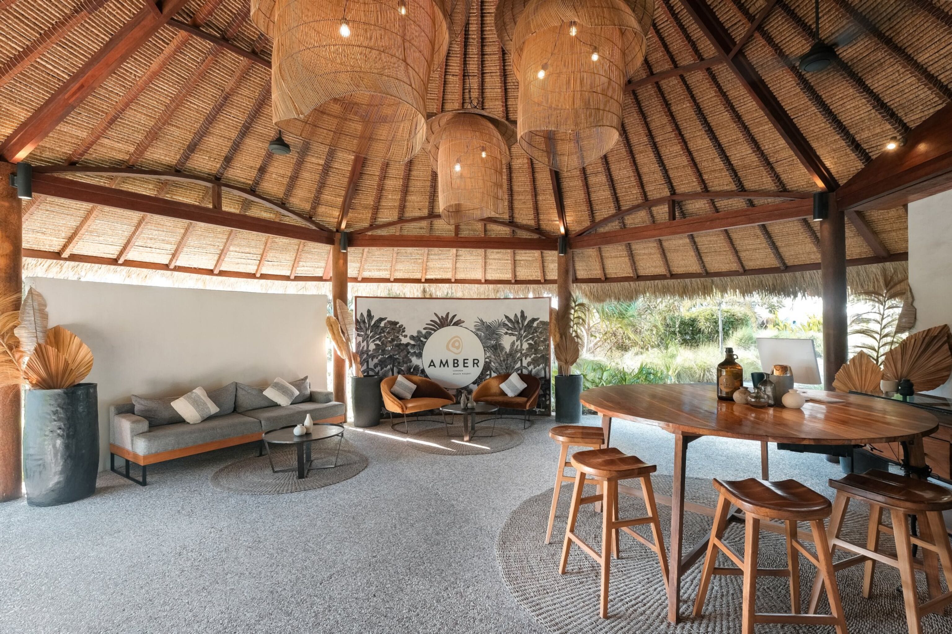Une pièce spacieuse avec un plafond très haut recouvert de chaume, un ensemble de canapés, une table à manger en bois avec des tabourets, et une grande fenêtre donnant sur un paysage verdoyant.
