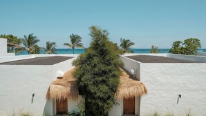 A white building with thatched roofs and palm trees, overlooking a clear blue sea.