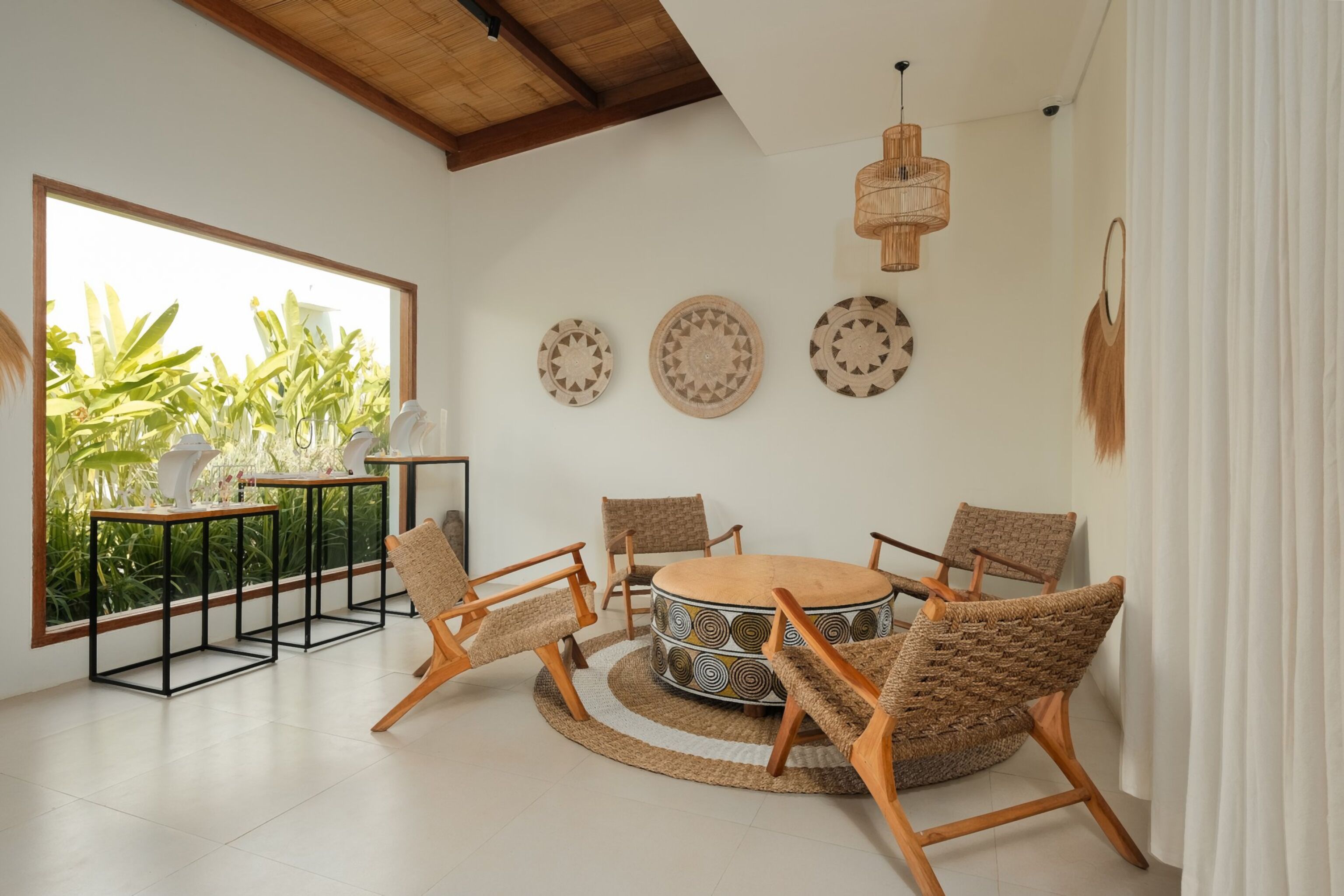 A modern living room with a round coffee table, four wicker chairs, a large window with plants, and decorative wall plates.